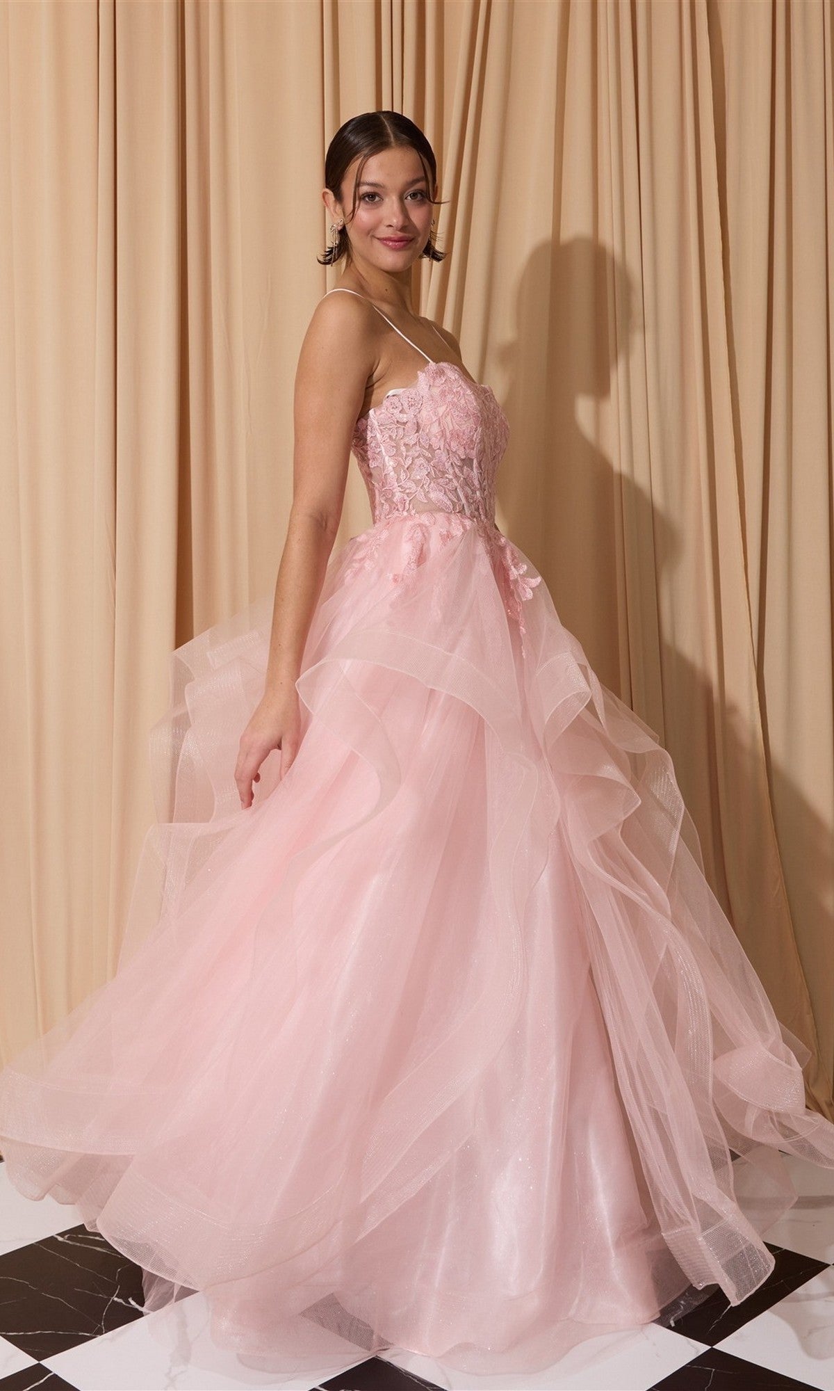 A woman poses in the Eureka EK167 long prom dress, a strapless light pink floral lace corset ball gown with a layered tulle skirt, standing on a black and white checkered floor before beige curtains.